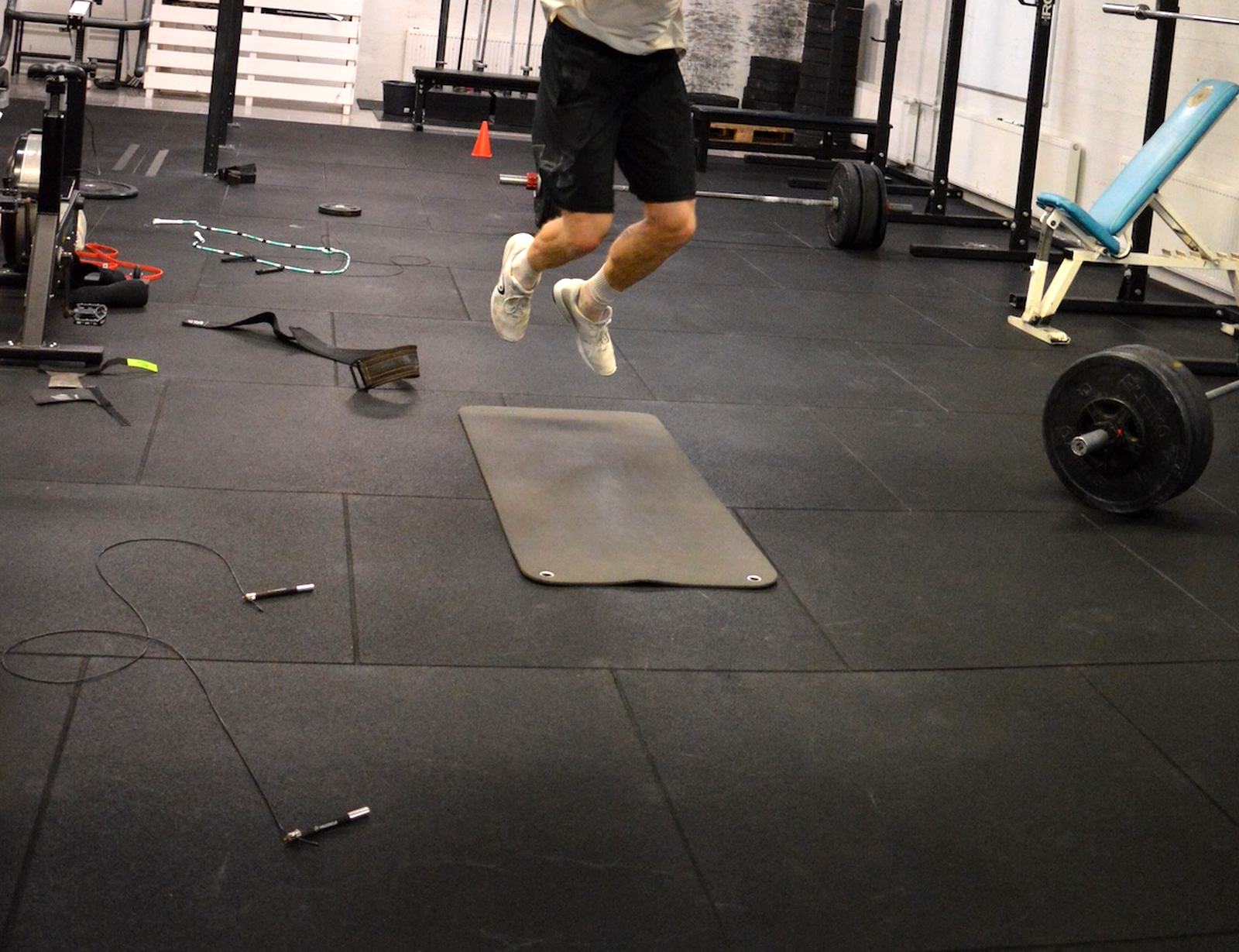 Treenaaja hypyn keskellä CrossFit SLN:n salilla, ympärillä levytanko, hyppynaruja ja patja